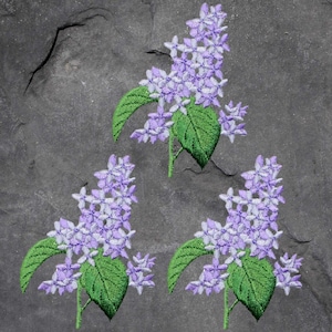 Könnte beinhalten: Drei gestickte Fliederblütenzweige mit grünen Blättern und Trauben kleiner lila Blüten. Die Stickerei ist detailliert, mit sichtbaren Stichen auf den Blättern und Blütenblättern. Der Hintergrund ist eine dunkelgraue, strukturierte Oberfläche.