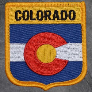 May include: Embroidered patch featuring the word "COLORADO" in black on a yellow background. Below, a blue field with a white stripe and a red and yellow circular emblem. The patch has a yellow border.