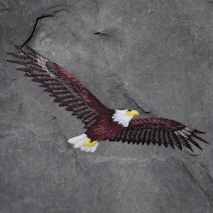 May include: An embroidered bald eagle in flight, with outstretched wings. The eagle features brown, white, and yellow details against a grey background. The bird's wings are spread wide, showcasing its detailed feathers.