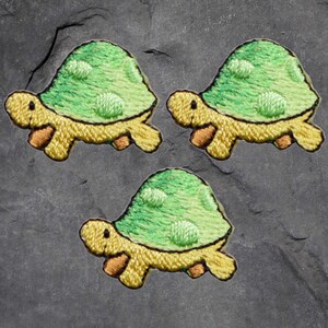 May include: Three embroidered turtle patches. Each patch features a green shell with lighter green spots, a yellow body, and brown feet. The patches are arranged on a dark gray, textured surface.