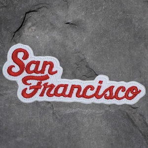 May include: Embroidered patch with the word "San Francisco" in red script lettering, outlined in white. The patch is rectangular and has a classic, vintage style. Perfect for jackets, bags, or other fabric items.