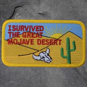 May include: Embroidered patch with the text "I Survived The Great Mojave Desert." The patch features a desert scene with a sun, mountains, a cactus, and a cow skull. The patch has a yellow border and a blue sky.