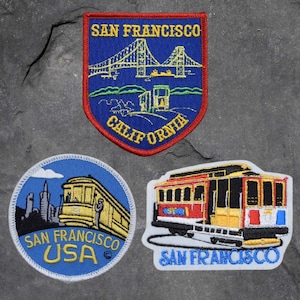 May include: Three embroidered patches featuring a San Francisco cable car and the Golden Gate Bridge. The patches are blue, yellow, and red with white stitching. The text on the patches reads "San Francisco, California, USA".