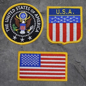 May include: Three embroidered patches with patriotic designs. One is a circular patch with the words "THE UNITED STATES OF AMERICA" and an eagle emblem. Another is a shield-shaped patch with "U.S.A." and red and white stripes. The third is a rectangular American flag.