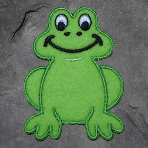 以下が含まれることがあります： 笑顔の顔と黒い刺繍が施された緑色のカエルのパッチ。カエルは、白と黒のアクセントが付いた大きくて丸い目を持っています。パッチは柔らかいフェルトのような素材で作られており、緑色のステッチで縁取られています。