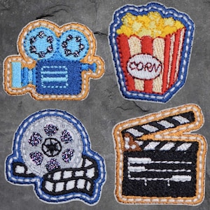 以下が含まれることがあります： 映画をテーマにしたデザインが施された刺繍パッチが4つ。パッチは、フィルムリール、映画カメラ、ポップコーンバケット、クラッパーボードの形をしています。パッチはすべて薄い青色の糸で縁取られており、白い背景になっています。