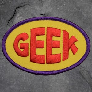 May include: Embroidered patch featuring the word "GEEK" in red, bold lettering. The text is set on a yellow oval, framed by a purple border. The patch is designed for application on clothing or accessories.