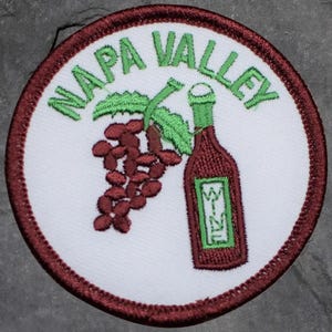 May include: Circular embroidered patch with the words "NAPA VALLEY" in green above a burgundy wine bottle and grapes. The bottle has a green label with "WINE". The grapes are deep red. The patch has a white background and a burgundy border.