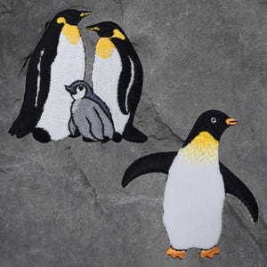 May include: Three embroidered penguin patches. Two adult penguins are standing side-by-side, with a smaller penguin chick standing between them. The adult penguins are black and white with yellow beaks and a yellow stripe around their eyes. The chick is grey with a black beak. The third penguin patch is a single penguin standing with its wings outstretched. It is black and white with a yellow beak and a yellow stripe around its eyes.