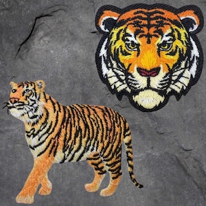 May include: Embroidered tiger patches in orange, black, and white. One patch depicts a full tiger, and the other shows a tiger's face. The patches are on a gray, textured surface.