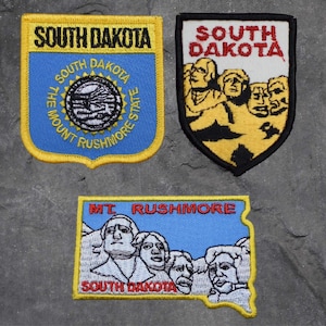 May include: Three embroidered patches featuring the state of South Dakota. The first patch is yellow with the text "South Dakota" in black and a blue background with the text "The Mount Rushmore State" in white. The second patch is yellow with the text "South Dakota" in red and white and features a black and white image of Mount Rushmore. The third patch is blue with the text "Mt. Rushmore" in red and white and features a black and white image of Mount Rushmore.