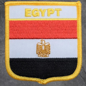 May include: Embroidered patch featuring the flag of Egypt. The patch has a white top section with the word "EGYPT" in gold letters, a red middle section, and a black bottom section. A gold emblem is in the white section.