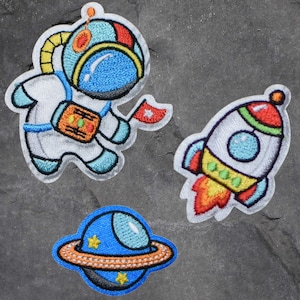 May include: Three embroidered iron-on patches featuring an astronaut, a rocket ship, and a planet. The astronaut is wearing a white spacesuit with a blue helmet and holding a red flag. The rocket ship is white with red, yellow, and green accents. The planet is blue with a yellow ring.