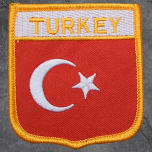 May include: Embroidered patch featuring the flag of Turkey. The patch has a red background with a white crescent moon and star. The word "TURKEY" is embroidered in yellow on a white banner at the top. The patch is bordered with yellow stitching.