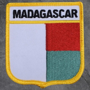 May include: Embroidered patch featuring the word "MADAGASCAR" above a shield design. The shield is divided into three sections: white, red, and green. The patch has a yellow border.