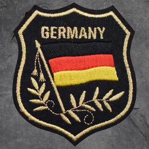 May include: Black embroidered patch with the word "GERMANY" above a flag in black, red, and yellow. The patch has a gold border and features a gold embroidered design of leaves and a flag pole.