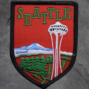 May include: Embroidered patch featuring the word "SEATTLE" in green letters above a red background. The patch depicts the Space Needle, a mountain range, and evergreen trees. The patch has a black border.