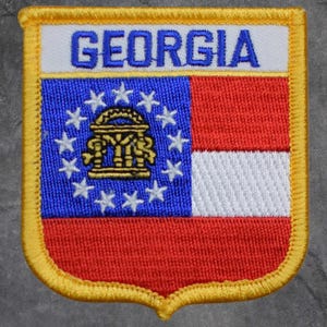 May include: Embroidered patch featuring the word "GEORGIA" above a design of the state flag. The patch has a blue field with white stars, a red and white striped section, and a gold border. The patch is rectangular with a curved bottom.