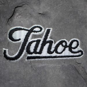 Könnte beinhalten: Gestickter Aufnäher mit dem Wort "Tahoe" in schwarzer Schrift mit weißer Umrandung. Der Aufnäher hat einen weißen Rand und ist vor einem grauen, strukturierten Hintergrund platziert.