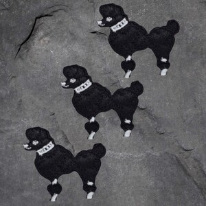May include: Three black felt poodle dog patches with white trim and a white collar. The patches are on a grey stone background.