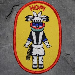 May include: Embroidered Hopi patch featuring a stylized figure with a yellow background and red border. The figure has a white headdress, black and white striped clothing, and is labeled "HOPI" in red text.