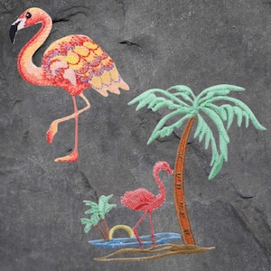 May include: Embroidered patch featuring two pink flamingos and a palm tree. The larger flamingo is standing on one leg, while the smaller flamingo is standing on both legs. The palm tree is green with a brown trunk.