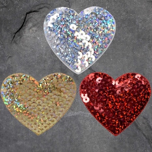 May include: Three heart-shaped decorations with sequin embellishments. One heart is clear with silver sequins, another is gold, and the third is red. The hearts are arranged on a dark gray surface.