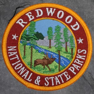 May include: Embroidered patch with the text "REDWOOD NATIONAL & STATE PARKS". The patch features a landscape scene with trees, a river, a deer, and a mountain range, all within a circular design with a gold border.