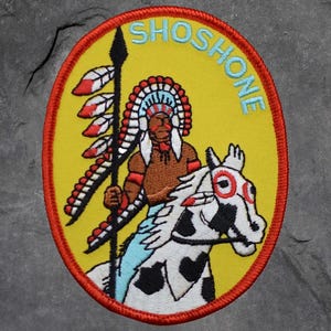 May include: Embroidered patch with a yellow background and an oval shape. The patch features a Native American figure riding a spotted horse, holding a spear. The word "SHOSHONE" is embroidered in light blue.