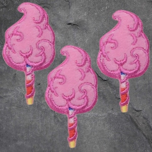 May include: Three embroidered patches of pink cotton candy on a stick. The cotton candy has a swirled design, and the stick is red and white striped with a yellow base. The patches are set against a grey background.