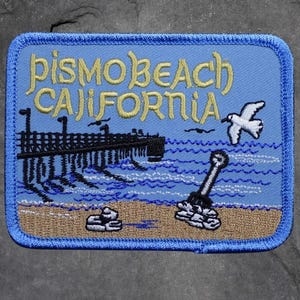 May include: A rectangular patch with a light blue background and a dark blue border. The patch features the words "Pismo Beach California" in yellow script. A pier, waves, a seagull, and other beach elements are also depicted.