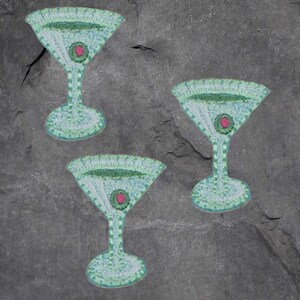 May include: Three embroidered patches in the shape of a martini glass. The patches are light blue with green detailing and a pink circular accent in the center. The patches are likely for clothing or accessories.