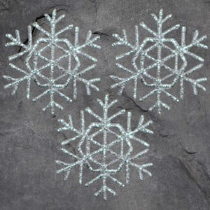 May include: Three silver snowflake ornaments with intricate designs are arranged on a dark gray, textured surface. The snowflakes have a delicate, sparkling appearance, suggesting a winter or holiday theme.