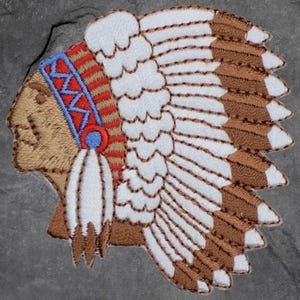 May include: Embroidered patch depicting a Native American headdress. The patch shows a profile with a tan face, a blue and red headband, and a large feathered headdress in white and brown. The design is detailed with intricate stitching.