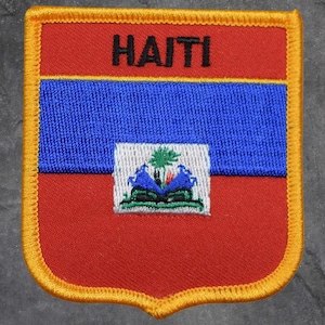 May include: Embroidered patch featuring the word "HAITI" in black letters above a blue stripe and a red background. The patch has a gold border and a central emblem with a palm tree and two blue mountains.