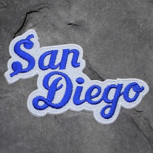 May include: Embroidered patch featuring the words "San Diego" in a stylized, cursive font. The text is in bright blue, outlined with white, and set against a white background. The patch is designed to be sewn or ironed onto fabric.