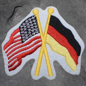 May include: Embroidered patch featuring the flags of the United States and Germany crossed. The American flag has red and white stripes with white stars on a blue field. The German flag has black, red, and yellow stripes.