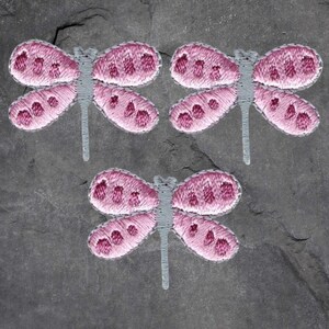 May include: Three embroidered dragonfly patches. Each dragonfly has pink wings with darker pink spots, a gray body, and a gray stem. The patches are on a dark gray, textured background.