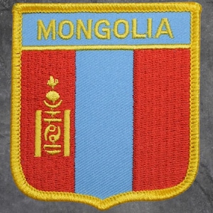 May include: Embroidered patch featuring the flag of Mongolia. The patch has a shield shape with a light blue stripe down the center, flanked by red stripes. The word "MONGOLIA" is embroidered in yellow at the top.