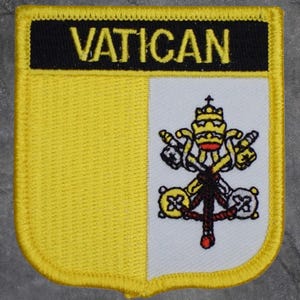 May include: A yellow and white Vatican patch with the word "VATICAN" in black block letters. The patch features the Vatican coat of arms, including a crown, keys, and a red cord.
