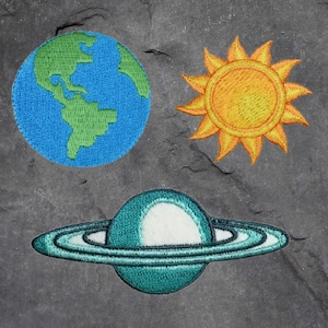 May include: Embroidered patches featuring a blue and green Earth, a yellow sun, and a teal planet with rings. The patches are designed with detailed stitching and vibrant colors, perfect for adding to clothing or accessories.