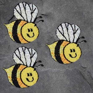 May include: Three embroidered bee patches. Each bee has a yellow body with black stripes, orange accents, and white wings. The bees have smiling faces and are set against a gray background.