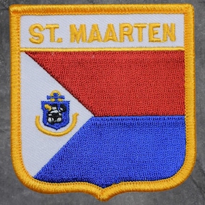 May include: Embroidered patch featuring the text "ST. MAARTEN" in gold lettering. The patch has a shield shape with a red, white, and blue design, and a gold border. A small emblem is on the white section.