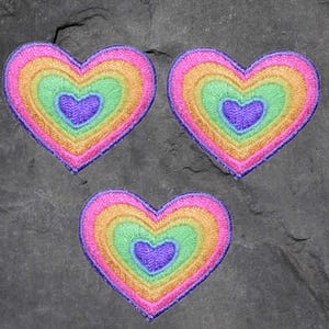 May include: Three embroidered heart patches in a rainbow of colors: pink, orange, yellow, green, and purple. The patches are layered, with each heart shape nested inside the other. The patches are on a dark gray, textured surface.