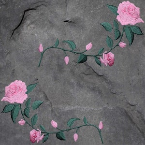 May include: Two pink roses with green leaves and stems are embroidered on a gray stone background.