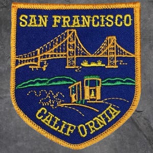 May include: Embroidered patch featuring the Golden Gate Bridge and a cable car, with the text "SAN FRANCISCO CALIFORNIA" in gold lettering on a blue background. The patch has a gold border.