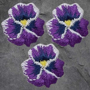 May include: Three embroidered pansy flower patches. Each patch features a vibrant purple petal design with a dark blue centre and a yellow centre. The edges are outlined with white stitching, creating a detailed and textured appearance.
