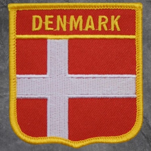 May include: A rectangular patch featuring the flag of Denmark. The patch has a red background with a white Scandinavian cross. The word "DENMARK" is embroidered in yellow at the top. The patch is outlined with a yellow border.