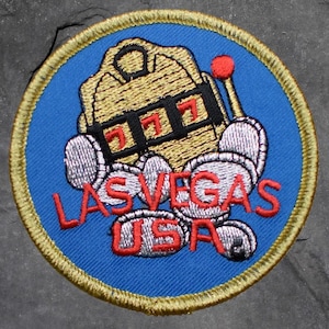 May include: Embroidered patch with a blue background and a gold border. The design features a gold slot machine with three red sevens, silver coins, and the words "LAS VEGAS USA" in red.