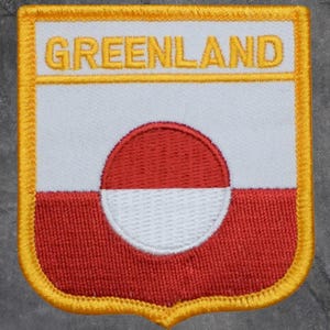 May include: Embroidered patch featuring the Greenland flag design. The patch has a white background with a red circle and a white half-circle. The bottom half is red. The word "GREENLAND" is embroidered in yellow.